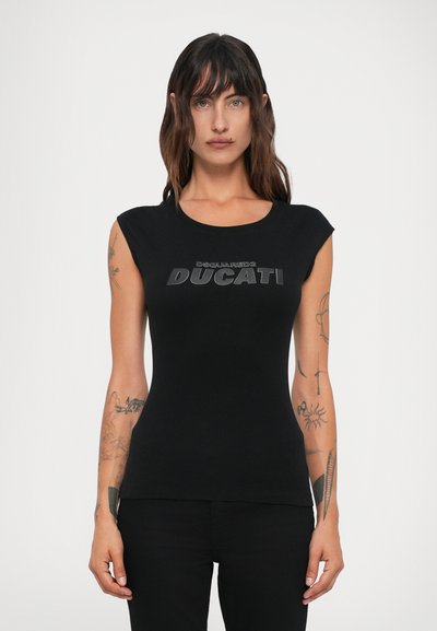 Donna con lunghi capelli scuri e tatuaggi su entrambe le braccia indossa un top aderente senza maniche nero con la scritta "DSQUARED2 DUCATI", abbinato a pantaloni neri.