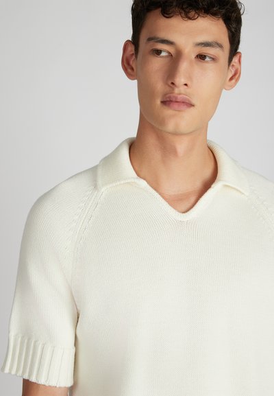 Giovane uomo con capelli corti e scuri che indossa una polo in maglia bianca a maniche corte, che guarda leggermente verso destra su uno sfondo semplice.