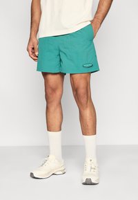 Τυρκουάζ μαγιό σορτς από ελαφρύ ύφασμα, με ελαστική μέση και λογότυπο στον αριστερό μηρό. Φορεμένα με λευκές κάλτσες και αθλητικά παπούτσια.