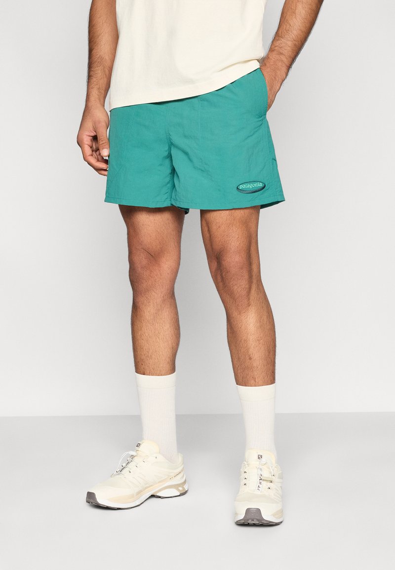 Τυρκουάζ μαγιό σορτς από ελαφρύ ύφασμα, με ελαστική μέση και λογότυπο στον αριστερό μηρό. Φορεμένα με λευκές κάλτσες και αθλητικά παπούτσια.