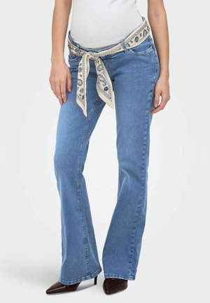 Naine kannab hele siniseid bootcut teksaseid, valget topi, mustrilist kangavööga vööd ümber piha ja pruune teravate ninadega kõrgete kontsadega kingi.