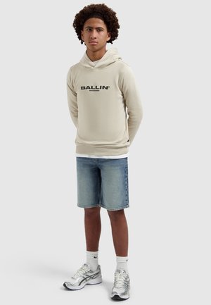 Teenager with curly hair wearing beige hoodie with "BALLIN'" text, denim shorts, white socks, and silver sneakers, standing with hands behind back.