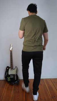 Bărbat purtând o cămașă verde măslinie și pantaloni negri, mergând departe de o chitară electrică alb-negru sprijinită de un perete alb, pe un podea de lemn.