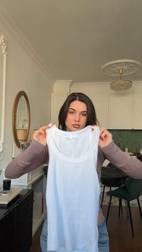 Bijela rebrasta majica bez rukava s okruglim izrezom; izrađena od mekanog materijala. U pozadini se vide tamni ormar i zelena zid.