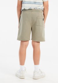 Olivgrüne Shorts mit weicher Textur, ausgestattet mit einer Gesäßtasche und einem elastischen Bund, kombiniert mit weißen Sneakern und Socken.