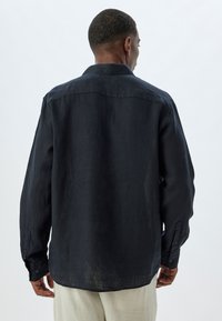 Camisa de lino azul oscuro con un corte relajado, mangas largas, puños con botones y un dobladillo curvado. Presenta una textura mínima y un cuello clásico.