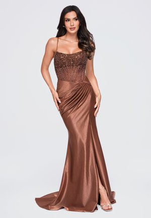 Femme aux longs cheveux foncés portant une robe en satin marron avec un corsage orné de perles et une fente latérale, posant les mains sur les hanches.