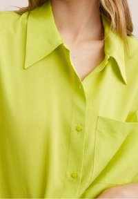 Camicia verde lime con colletto a punta, maniche corte e taschino sul petto. Tessuto morbido con due bottoni gialli.