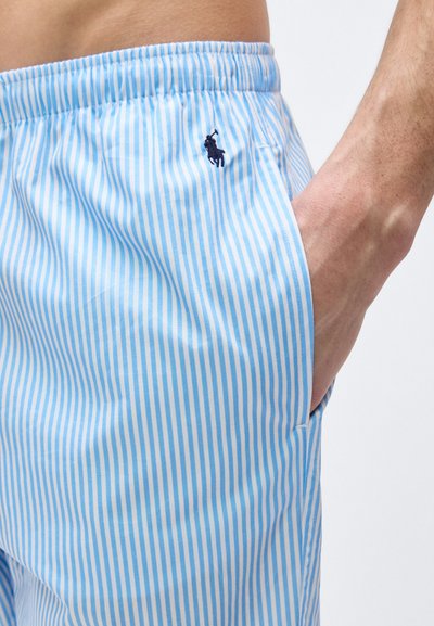 Κοντινή εικόνα του χεριού ενός ατόμου στην τσέπη ενός ζευγαριού σορτς με ανοιχτό μπλε και λευκές ρίγες, με ελαστική μέση και μικρό σκούρο λογότυπο.