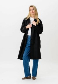 Femme blonde portant un long manteau noir par-dessus une chemise blanche et un jean bleu, debout devant un fond blanc uni.