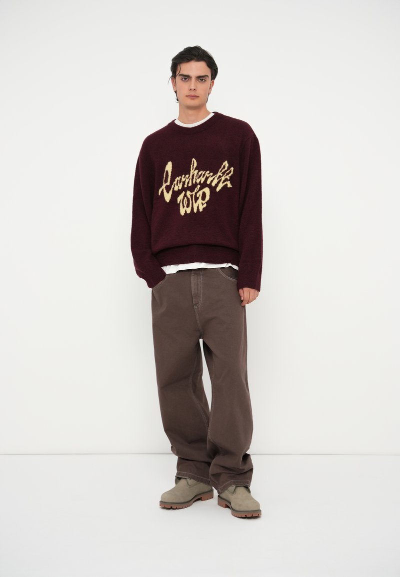 Bordeaux gebreide trui met gouden "Carhartt WIP" letters, gecombineerd met losse bruine broek en grijze laarzen. Eenvoudige, casual outfit tegen een witte achtergrond.