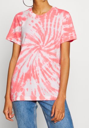 Osoba nosząca koralowo-biały t-shirt krótkim rękawem z wirowym tie-dye w połączeniu z niebieskimi dżinsami, stojąca na jednolitym tle.
