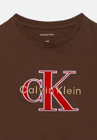Καφέ βαμβακερό T-shirt με μεγάλο κεντημένο λογότυπο "CK" σε κόκκινο και χρυσό, με το κείμενο "Calvin Klein" από κάτω σε μικρότερη χρυσή γραμματοσειρά.