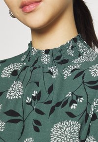 Grön blommig blus med elastisk krage, med vita och svarta blommönster på ett strukturerat tyg.