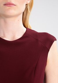 Bordeaux mouwloze jurk met een hoge halslijn en gestructureerde schouders, gemaakt van een gladde, textuurstof met naaddetails.