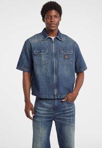 Giubbotto di jeans blu scuro, maniche corte, chiusura a zip, due tasche sul petto e una patch con il marchio. Abbinato a pantaloni in jeans coordinati.