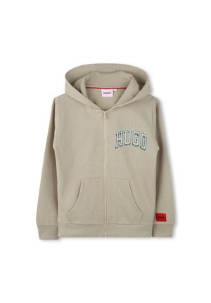 Un sweat à capuche zippé beige en tissu doux. Présente un grand logo vert "HUGO", deux poches avant et une étiquette rouge en bas à droite.