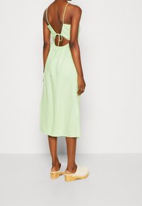Hellgrünes Leinenkleid mit dünnen Trägern, offenem Rücken und einem Bindedetail, knielang. Kombiniert mit beigen Holzclogs.