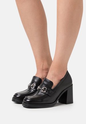 Mocassins en cuir noir avec un bout arrondi, dotés d'une bride décorative à boucle en argent et d'un talon épais noir. Texture lisse avec un minimum de couture.
