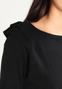 Zwarte trui met geribbelde textuur, voorzien van decoratieve ruches op de schouders en een brede halslijn.