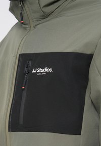 Giacca verde oliva con tasca sul petto nera con zip e dettaglio logo bianco. Tessuto liscio con blocchi di colore a contrasto.