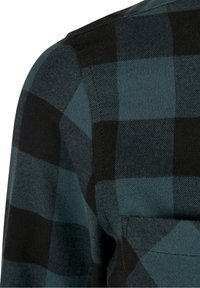 Gros plan d'une chemise en tissu à carreaux vert foncé et noir montrant la partie supérieure de la manche et une partie de la poche poitrine.