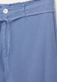 Pantalon en lin bleu avec un bouton, ceinture en tissu nouée à la taille et bord de ceinture effiloché.