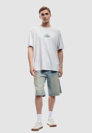 Camiseta blanca de algodón con gráfico "FORE!" en verde, combinada con pantalones cortos de mezclilla azul claro y zapatillas blancas. Fondo liso.
