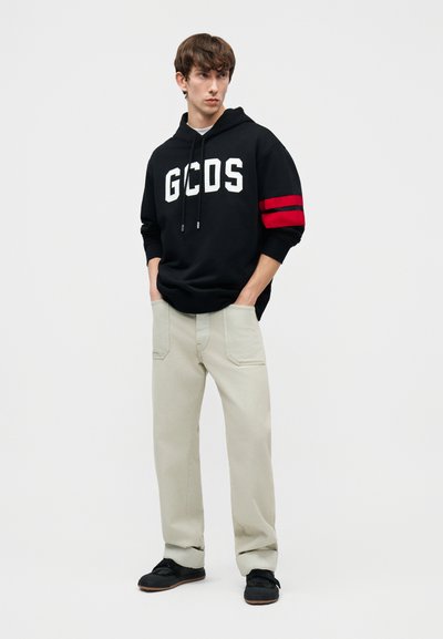 Ung mann som har på seg en svart GCDS-hettegenser med røde striper på venstre arm og lyse beige bukser, står med hendene i lommene.