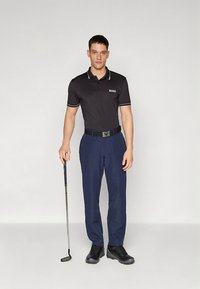 Fekete galléros póló világosbarna részletekkel, tengerészkék nadrág, fekete öv és sötét cipő; a modell golfütőt tart, egyszerű háttér előtt áll.