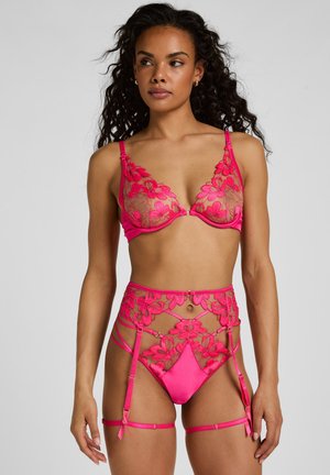 Femme portant un ensemble de lingerie en dentelle florale rose comprenant un soutien-gorge, une jarretière taille haute et une culotte assortie, debout devant un fond uni.