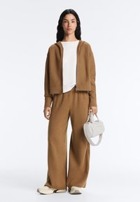 Femme portant un sweat à capuche zippé marron à texture et un pantalon large assorti, chemise blanche, baskets crème, tenant un petit sac à main blanc.