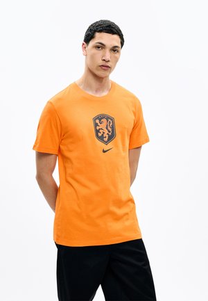 Jonge man die een oranje Nike T-shirt draagt met een zwart leeuwenwapen en zwarte broek, staand met de handen achter de rug tegen een witte achtergrond.