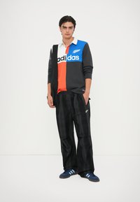Črne karirane hlače, dolgi rokavi s kratkim ovratnikom v modri, oranžni in sivi barvi z logotipom. Model nosi temno modresuperge s tremi črtami.
