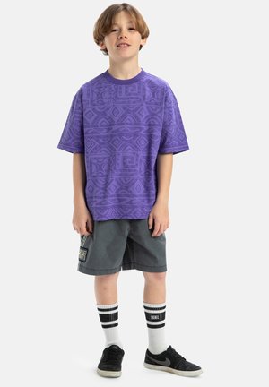Paarse kortemouwen shirt met geometrisch patroon, grijze shorts, zwarte sneakers, witte sokken met zwarte strepen, staande pose.