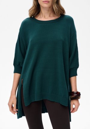 Kobieta w luźnym, ciemnozielonym swetrze z długim rękawem do trzech kwart, z bocznymi rozcięciami, w połączeniu z ciemnymi legginsami i bransoletkami.