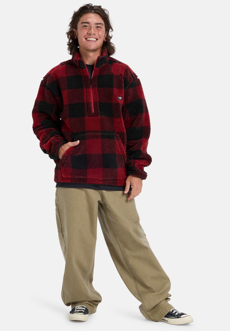 Red and black plaid fleece pullover with a quarter zip. Paired with loose beige pants and black sneakers, showcasing a relaxed fit.
