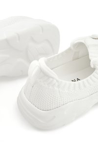 White slip-on sneakers with a textured knit upper, padded collar, and a thick, flexible rubber sole featuring curved treads.