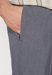 Hand in zijzak van grijze broek gestoken met zichtbare rits en daarboven een beige gebreide mouwrand van een trui.