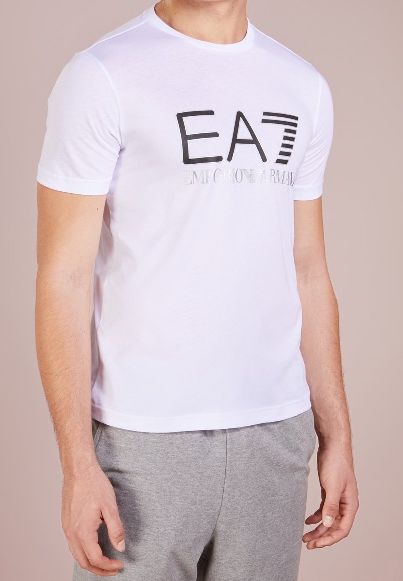 Biała bawełniana koszulka z krótkim rękawem, z czarnym nadrukowanym logo "EA7" oraz "EMPORIO ARMANI" z przodu. Gładka struktura.