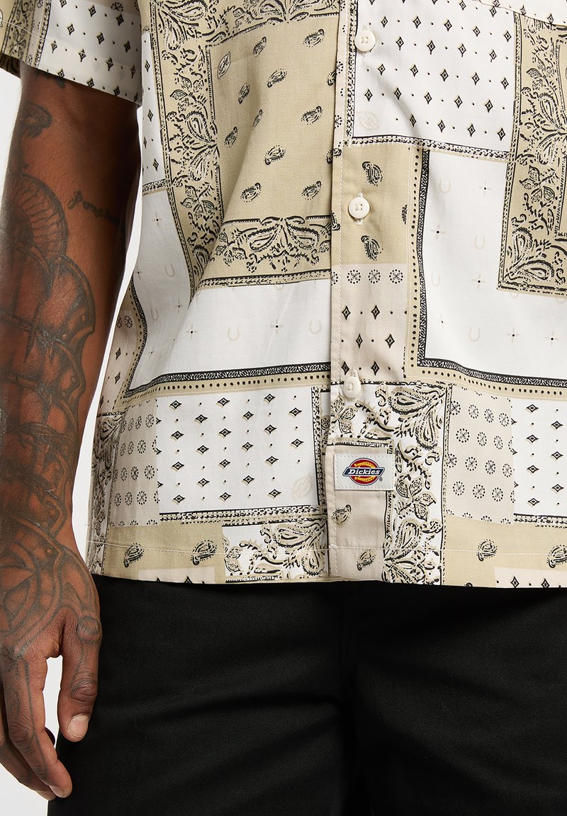 Chemise à manches courtes beige et blanche, ornée de motifs variés en cachemire et losanges. Détail notable : étiquette Dickies sur l'ourlet.