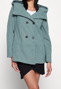 Teal wolblend dubbelzijdig jasje met een afneembare capuchon, voorkeurszakken en zwarte knopen. Getextureerde stof met een subtiel visgraatpatroon.