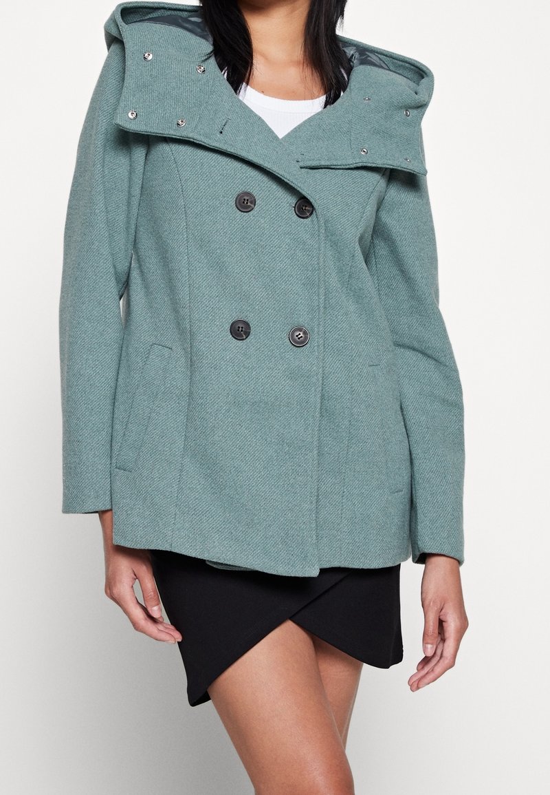 Teal wolblend dubbelzijdig jasje met een afneembare capuchon, voorkeurszakken en zwarte knopen. Getextureerde stof met een subtiel visgraatpatroon.