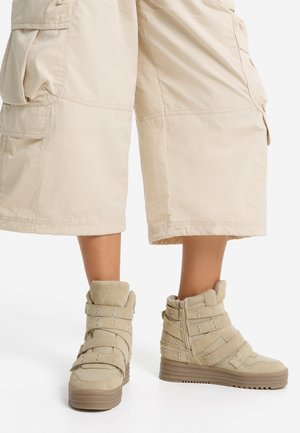 Gros plan d'un mannequin portant un short cargo crème et des bottes montantes beiges avec des brides cloutées et des semelles épaisses.