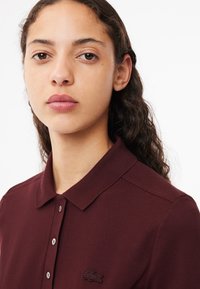 Polo bordeaux con colletto classico, caratterizzato da un tessuto testurizzato, una chiusura a tre bottoni e un logo ricamato nero sul petto.