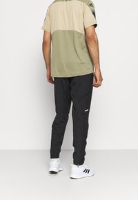 Olivgrünes und beiges Sportshirt kombiniert mit schwarzen, schmal zulaufenden Hosen. Merkmale sind kurze Ärmel und reflektierende Akzente. Weiße Sportschuhe.