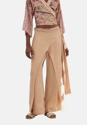 Pantalones envolventes beige con una textura suave y piernas anchas, que presentan un cinturón de lazo y borlas decorativas en el dobladillo. Combinados con un top corto con estampado.