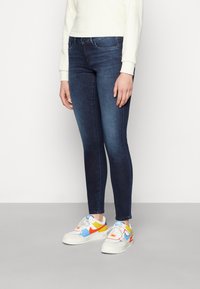 Persona che indossa jeans skinny blu scuro, felpa bianca e sneakers bianche con accenti blu, gialli e arancioni, in piedi su un pavimento grigio.