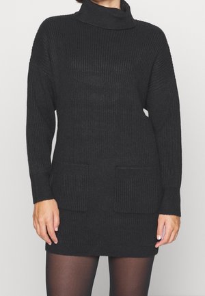 Robe pull en maille côtelée noire avec un col montant, des manches longues et deux poches avant, présentant un tissu texturé et une coupe décontractée.