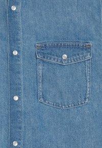 Svetle modrá denimová košile s knoflíkovým zapínáním vpředu, zdobená stříbrnými cvoky a kapsou na prsou s kontrastním švem.